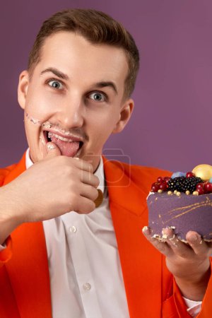 Photo for Handsome birthday man in orange jacket eating cream cake on purple background - Royalty Free Image