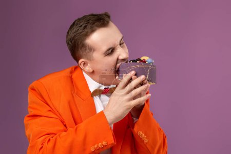 Photo for Birthday man in orange jacket bites cake on purple background. copy space - Royalty Free Image