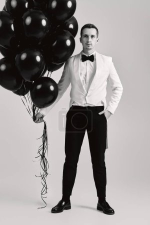 Téléchargez les photos : Élégant homme caucasien en costume blanc smoking avec ballons à air noir sur fond blanc. - en image libre de droit
