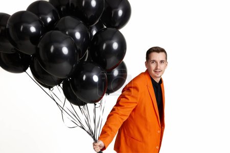 Téléchargez les photos : Homme heureux en veste orange avec des ballons à air noir sur fond blanc. fête d'anniversaire - en image libre de droit