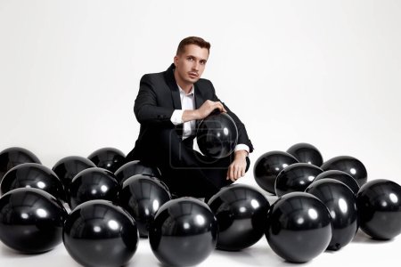 Foto de Elegante hombre caucásico en traje esmoquin sentado en el piso del estudio con una gran cantidad de globos de aire negro - Imagen libre de derechos