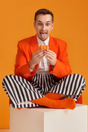 Téléchargez les photos : Jeune homme drôle extravagant en veste orange et pantalon rayé tenant cupcake sur fond orange. - en image libre de droit