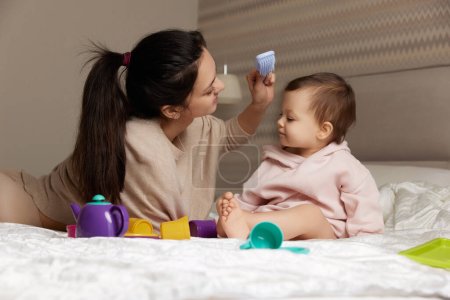 Téléchargez les photos : Belle mère peigne ses mignons cheveux de petite fille sur le lit - en image libre de droit