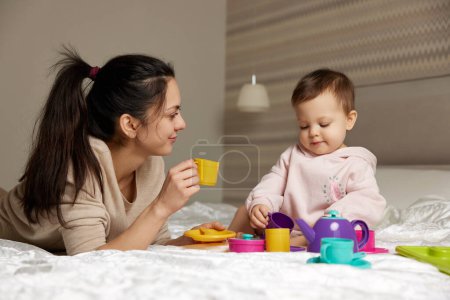 Foto de Feliz madre e hija pequeña fingiendo beber té de vasos de juguete de plástico y pasar tiempo juntos en el dormitorio - Imagen libre de derechos