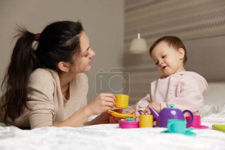 Téléchargez les photos : Mère heureuse et petite fille enfant prétendant boire du thé à partir de tasses à jouets et passer du temps ensemble dans la chambre à coucher, la famille s'amuse - en image libre de droit