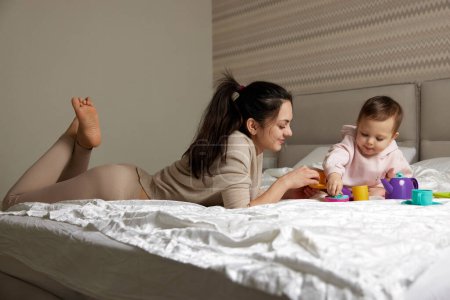 Téléchargez les photos : Mère heureuse et petite fille enfant jouant thé partie et passer du temps ensemble sur le lit - en image libre de droit