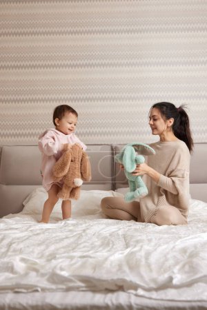 glückliche Mutter und ihre kleine Tochter spielen mit Hasenspielzeug im Schlafzimmer, Familie hat Spaß
