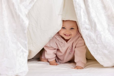 Foto de Linda niña divertida mira hacia fuera desde debajo de la manta en la cama, espacio de copia - Imagen libre de derechos