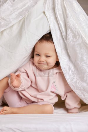 Téléchargez les photos : Mignonne petite fille drôle regarde dehors sous la couverture sur le lit - en image libre de droit