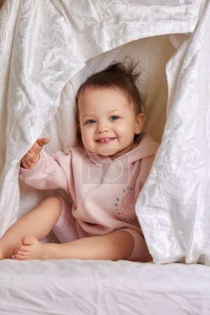 Téléchargez les photos : Mignonne fille drôle bébé caché sous la couverture sur le lit - en image libre de droit