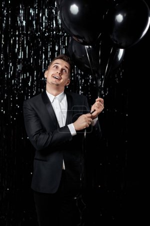 Foto de Hombre guapo feliz en chaqueta negra sosteniendo globos de aire negro sobre fondo de brillo. - Imagen libre de derechos