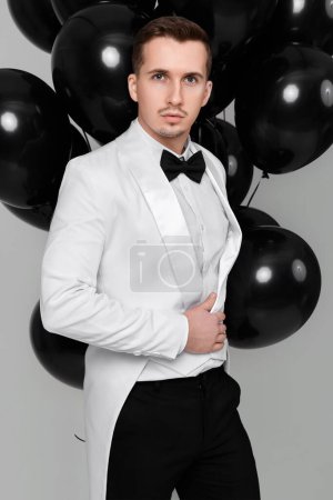Téléchargez les photos : Élégant homme caucasien en costume blanc smoking avec bouquet de ballons à air noir sur fond blanc. célébration des fêtes - en image libre de droit