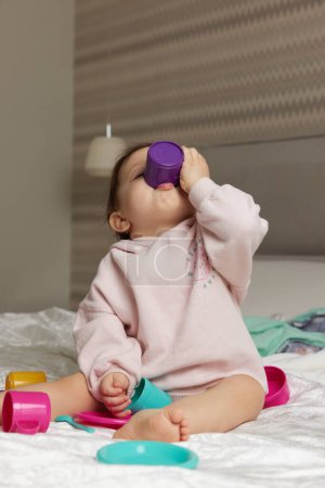 Foto de Feliz linda niña jugando y fingiendo beber té de tazas de juguete en la cama - Imagen libre de derechos