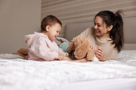 Foto de Feliz madre y su hija pequeña jugando con juguetes de conejito en el dormitorio, la familia se divierte - Imagen libre de derechos