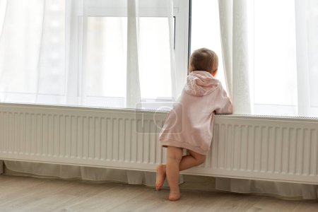 Photo for Little child girl standing at window and looking outside. Back view. - Royalty Free Image