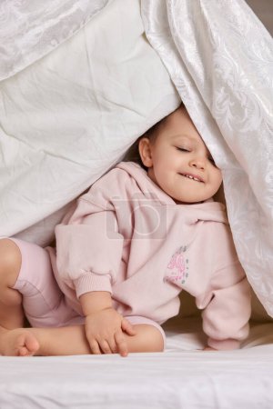 Téléchargez les photos : Mignonne fille drôle bébé caché sous la couverture sur le lit - en image libre de droit