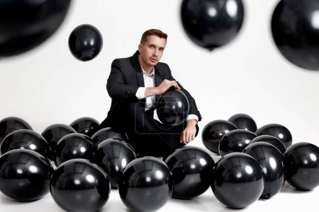 Foto de Elegante hombre caucásico en traje esmoquin sentado en el piso del estudio con una gran cantidad de globos de aire negro - Imagen libre de derechos