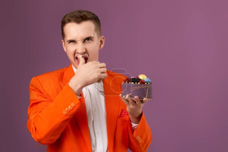 Photo for Handsome birthday man in orange jacket eating cake on purple background - Royalty Free Image