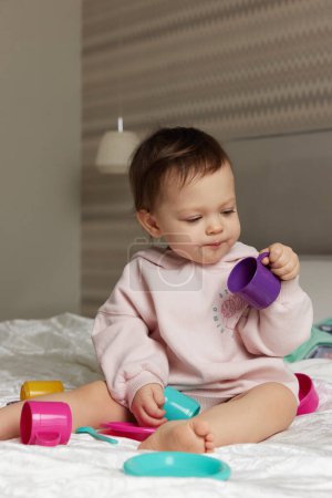 Téléchargez les photos : Heureux mignon petit enfant fille jouer et faire semblant de boire du thé à partir de tasses jouet sur le lit - en image libre de droit