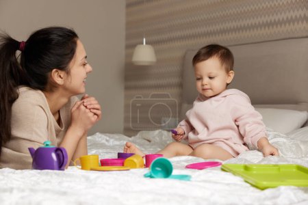Foto de Sonriente madre e hija pequeña jugando fiesta de té y pasar tiempo juntos en el dormitorio, la familia se divierten - Imagen libre de derechos