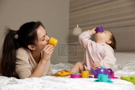 Foto de Feliz madre e hija pequeña fingiendo beber té de vasos de juguete de plástico en el dormitorio, la familia se divierte - Imagen libre de derechos