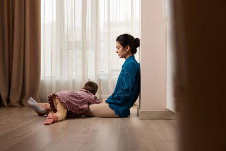 Téléchargez les photos : Femme fatiguée assise sur le sol avec sa petite fille à la maison - en image libre de droit