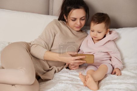 Téléchargez les photos : Mère et enfant caucasiens jouant avec smartphone assis sur le lit dans la chambre à coucher à la maison - en image libre de droit
