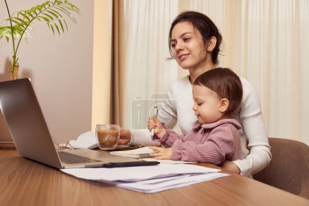 Photo for Cheerful pretty businesswoman working on laptop at home with her little child girl. mom spending time with her cute baby - Royalty Free Image