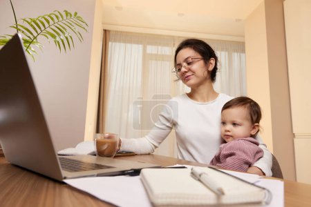 Foto de Alegre bastante mujer de negocios trabajando en el ordenador portátil en casa con su niña - Imagen libre de derechos