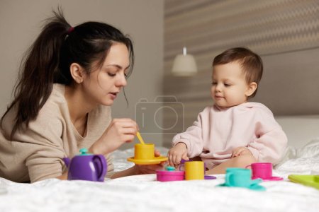 Photo for Attractive mother and little child daughter playing tea party and spending time together in bedroom, family having fun - Royalty Free Image