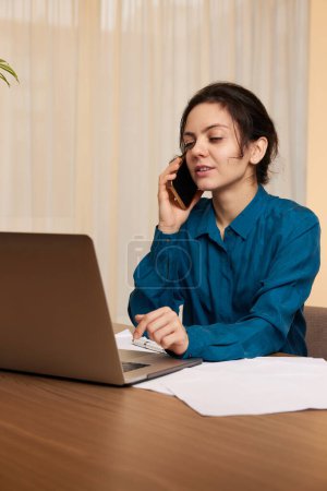 Foto de Hermosa mujer de negocios que trabaja en el ordenador portátil y se comunica con el cliente por teléfono en casa - Imagen libre de derechos