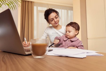 Photo for Tired woman working on laptop at home with her little baby girl. Child makes noise and disturb mother at work. - Royalty Free Image