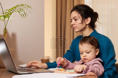 Photo for Beautiful businesswoman working on laptop with her little child girl at home - Royalty Free Image