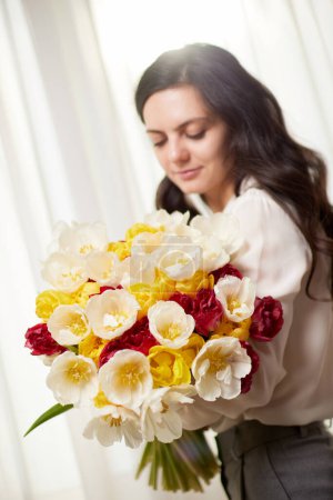 Téléchargez les photos : Belle femme dans la chemise blanche avec des tulipes de fleurs de printemps dans les mains. Journée de la femme. accent sur la fleur - en image libre de droit