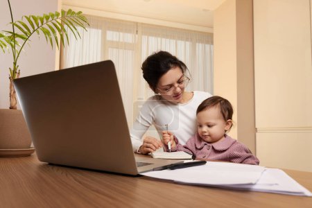 Téléchargez les photos : Femme d'affaires positive dans des lunettes de travail sur ordinateur portable à la maison avec sa petite fille enfant. Reste à la maison. - en image libre de droit