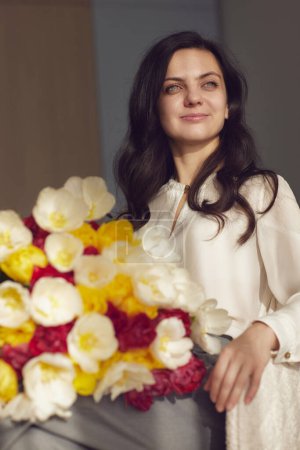Photo for Beautiful woman with spring flowers tulips in hands sitting on chair near window. Womens Day - Royalty Free Image