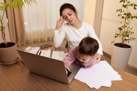 Foto de Mujer estresada trabajando en la computadora portátil en casa con su pequeña niña. El niño hace ruido y molesta a la madre en el trabajo. - Imagen libre de derechos