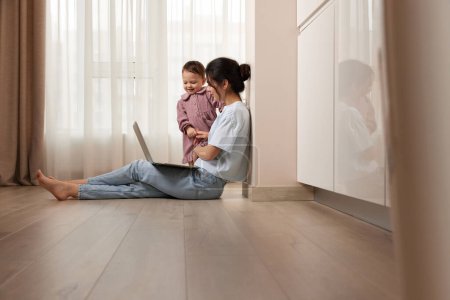 Foto de Hermosa mujer casual sentada en el suelo y trabajando en el ordenador portátil con su pequeña niña en casa - Imagen libre de derechos