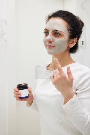 Téléchargez les photos : Femme caucasienne regardant dans le miroir et appliquant masque blanc dans la salle de bain - en image libre de droit