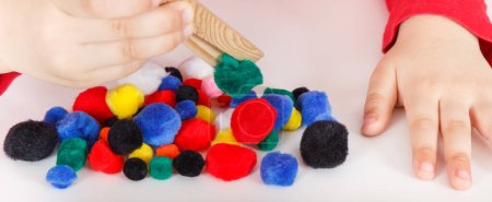 Little baby boy playing with small fluffy colorful pompoms and wooden tongs. Development of kids motor skills, coordination, creativity and logical thinking