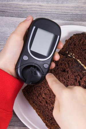 Téléchargez les photos : Main d'enfant tenant glucomètre, gâteau au chocolat sucré. Mesurer et contrôler le taux de sucre pendant le diabète - en image libre de droit