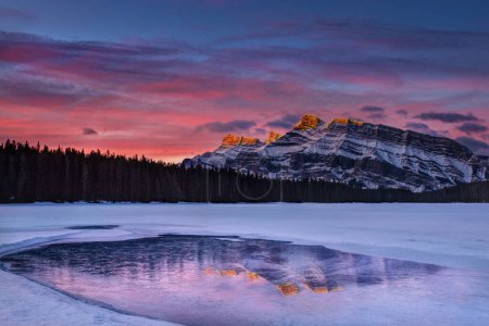 Goldener Sonnenaufgang am Two Jack Lake mit Alpenglühen auf den Gipfeln des Mount Rundle, die sich am zugefrorenen See mit Schnee und Eis spiegeln. 