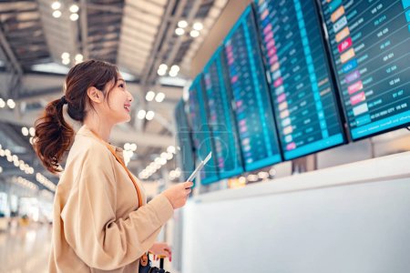 Photo for Happy asian woman traveller checking flight schedule departures board in airport terminal hall in front of check in counters. Tourist journey trip concept - Royalty Free Image