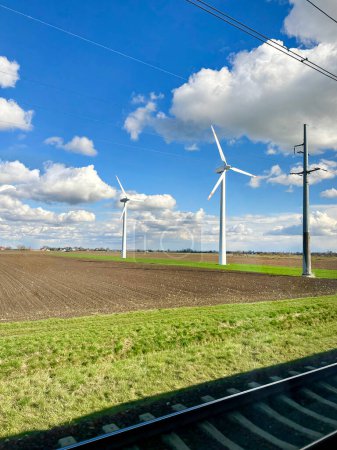 Landwirtschaftliches Feld umgeben von Windmühlen mit hohen Windrädern zur Stromerzeugung mit Stromleitungen und Eisenbahnen. Grünes Energiekonzept. Windenergieproduktion