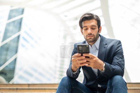 Foto de Primer plano de un hombre joven caucásico serio usando un dispositivo de teléfono inteligente moderno mientras está sentado en una zona pública, mientras mira la red social en su teléfono inteligente. - Imagen libre de derechos