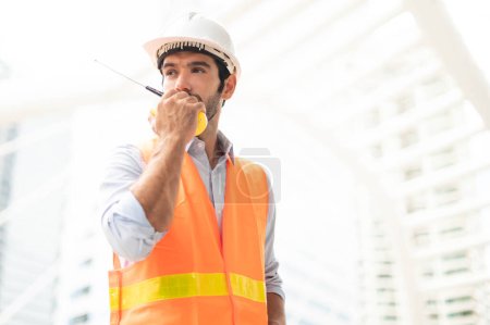 Foto de Ingenieros caucásicos usan un walkie-talkie para hablar, con chalecos anaranjados y grandes sombreros duros en el trabajo del sitio del centro de la ciudad. - Imagen libre de derechos