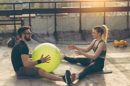 Photo for Couple sitting on a yoga mat on a building rooftop terrace, exercising with a pilates ball - Royalty Free Image