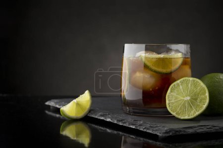 Téléchargez les photos : Cuba Cocktail libre au rhum brun, jus de citron, coke et glaçons, décoré de tranches de lime fraîche sur un plateau moderne en pierre noire - en image libre de droit