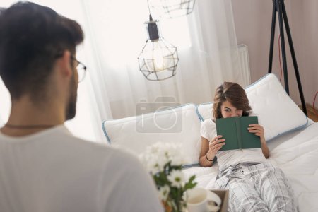 Téléchargez les photos : Joyeux et surpris jeune femme couchée dans le lit le matin tandis que son mari apporte son petit déjeuner et des fleurs sur un plateau - en image libre de droit