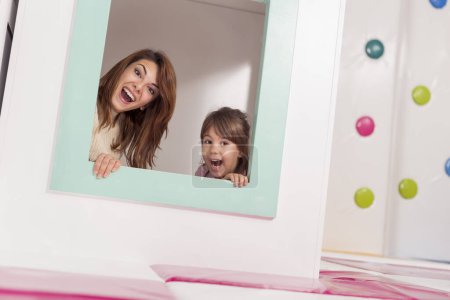 Photo for Cheerful mother and daughter hiding in a small wooden house in a playroom, peeking through a window and making faces - Royalty Free Image